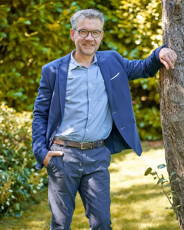 Dr. Theissing steht in einem sonnigen Garten, eine Hand in der Hosentasche, ein Arm locker an einem Baum gestützt.