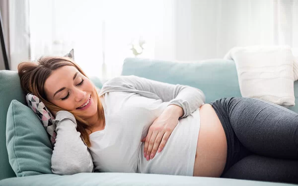 Schwangere Frau liegt lächelnd auf der Couch und streichelt ihren Bauch.