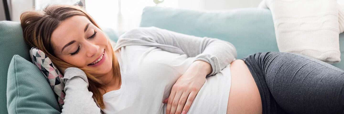 Schwangere Frau liegt lächelnd auf der Couch und streichelt ihren Bauch.