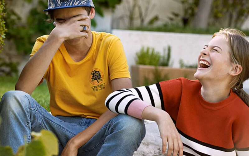Ein Junge und ein Mädchen sitzen nebeneinander in einem Park und lachen ausgelassen.