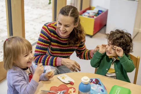 Zwei Kinder sitzen mit einer Erzieherin lachend an einem Tisch und essen ihr Brot. Das Kita-Programm JolinchenKids der AOK fördert die Gesundheit von Kindern.