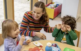 Zwei Kinder sitzen mit einer Erzieherin lachend an einem Tisch und essen ihr Brot. Das Kita-Programm JolinchenKids der AOK fördert die Gesundheit von Kindern.