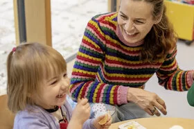 Zwei Kinder sitzen mit einer Erzieherin lachend an einem Tisch und essen ihr Brot. Das Kita-Programm JolinchenKids der AOK fördert die Gesundheit von Kindern.