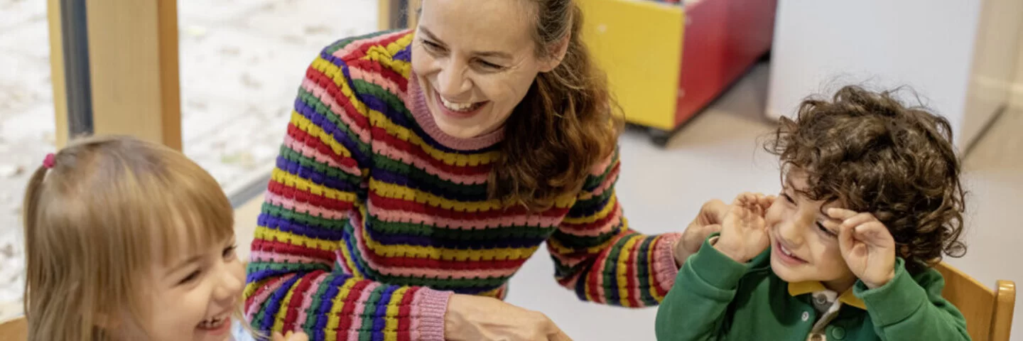 Zwei Kinder sitzen mit einer Erzieherin lachend an einem Tisch und essen ihr Brot. Das Kita-Programm JolinchenKids der AOK fördert die Gesundheit von Kindern.