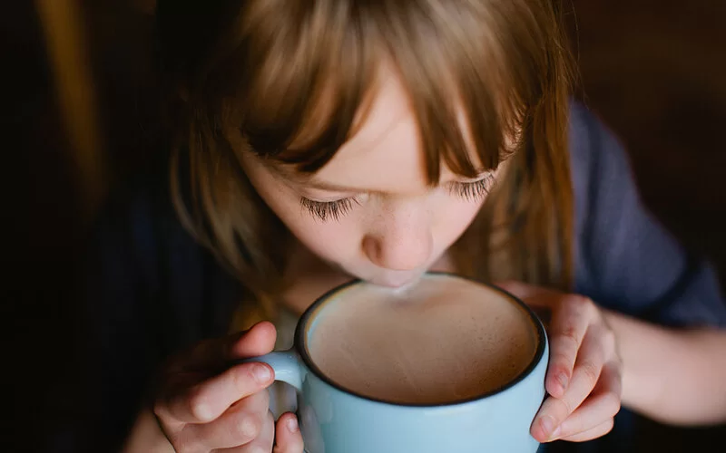 Ein Mädchen sitzt am Esstisch und hält mit beiden Händen eine große Tasse warmen Kakao, aus der sie vorsichtig trinkt.