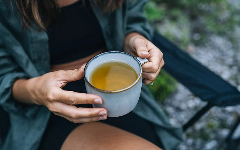 Eine junge Frau sitzt draußen und hält eine Keramiktasse randvoll mit Tee.