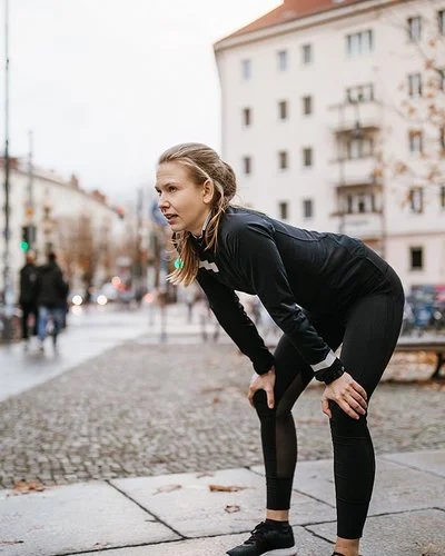 Eine junge Frau in Laufkleidung und urbaner Umgebung wirkt erschöpft. Sie beugt sich vor und stützt die Hände auf den Knien ab.