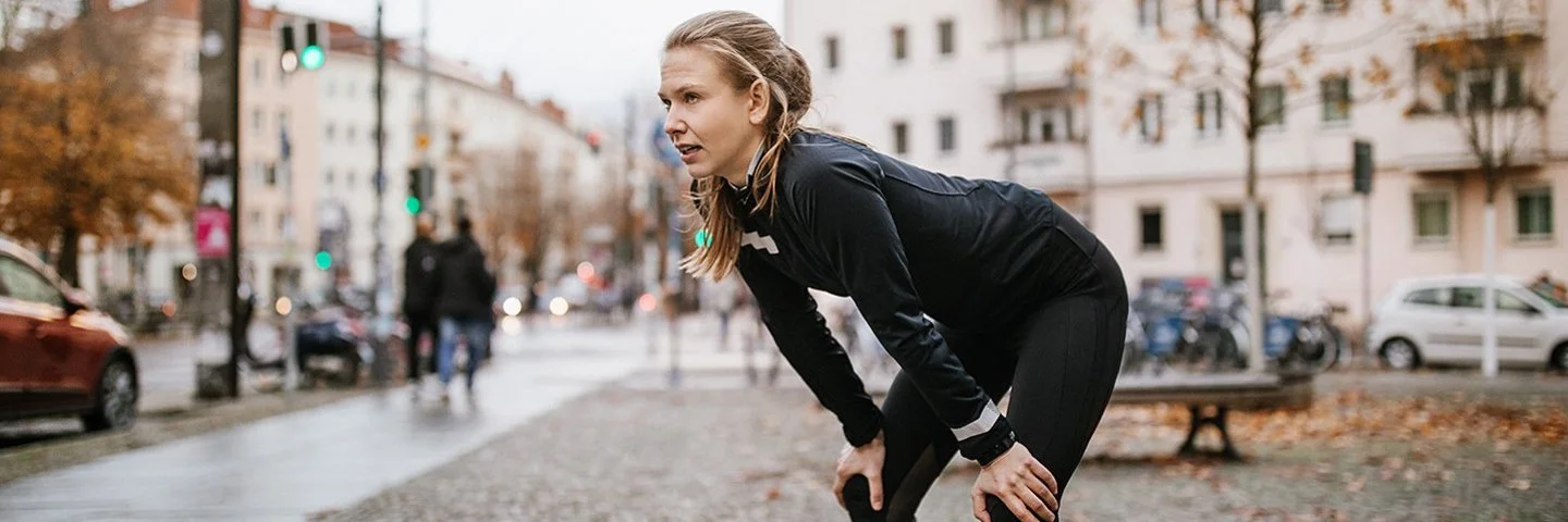 Eine junge Frau in Laufkleidung und urbaner Umgebung wirkt erschöpft. Sie beugt sich vor und stützt die Hände auf den Knien ab.