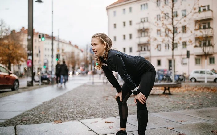 Eine junge Frau in Laufkleidung und urbaner Umgebung wirkt erschöpft. Sie beugt sich vor und stützt die Hände auf den Knien ab.