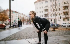 Eine junge Frau in Laufkleidung und urbaner Umgebung wirkt erschöpft. Sie beugt sich vor und stützt die Hände auf den Knien ab.