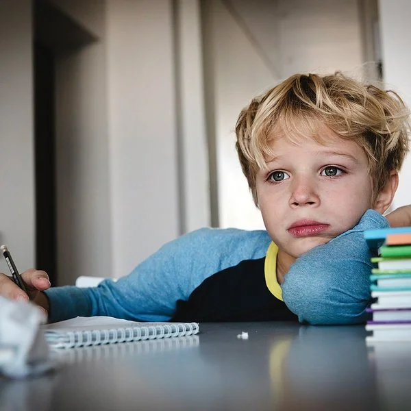 Ein Schuljunge sitzt an einem Tisch. Auf dem Tisch liegen ein Schreibblock, ein Stapel Lernmaterialien sowie zerknüllte Papierkugeln. In seiner rechten Hand hält er einen Stift. Er blickt nicht auf den Block, sondern nach vorne.