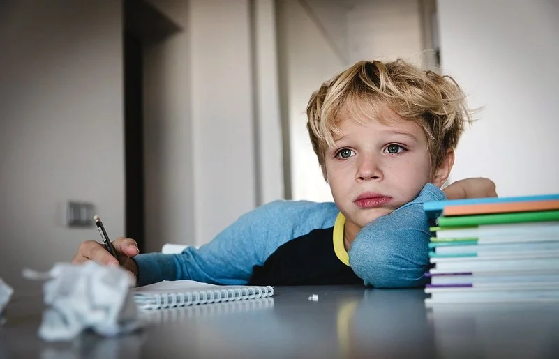 Ein Schuljunge sitzt an einem Tisch. Auf dem Tisch liegen ein Schreibblock, ein Stapel Lernmaterialien sowie zerknüllte Papierkugeln. In seiner rechten Hand hält er einen Stift. Er blickt nicht auf den Block, sondern nach vorne.