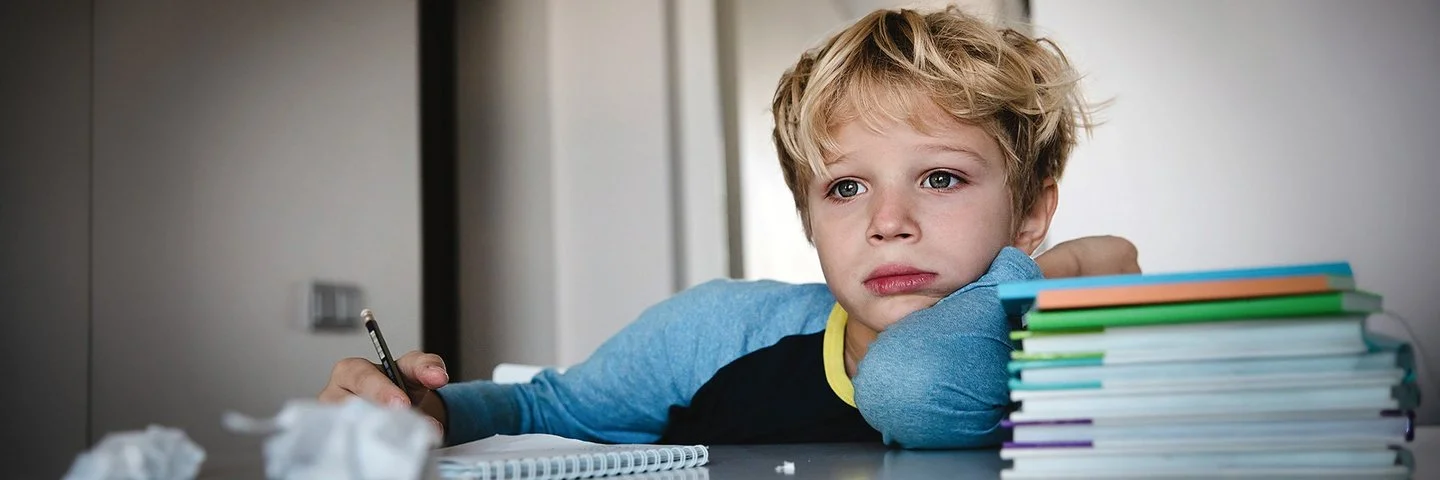 Ein Schuljunge sitzt an einem Tisch. Auf dem Tisch liegen ein Schreibblock, ein Stapel Lernmaterialien sowie zerknüllte Papierkugeln. In seiner rechten Hand hält er einen Stift. Er blickt nicht auf den Block, sondern nach vorne.