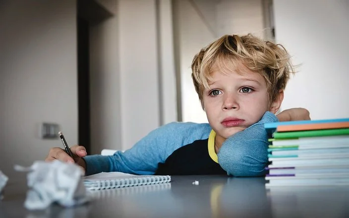 Ein Schuljunge sitzt an einem Tisch. Auf dem Tisch liegen ein Schreibblock, ein Stapel Lernmaterialien sowie zerknüllte Papierkugeln. In seiner rechten Hand hält er einen Stift. Er blickt nicht auf den Block, sondern nach vorne.