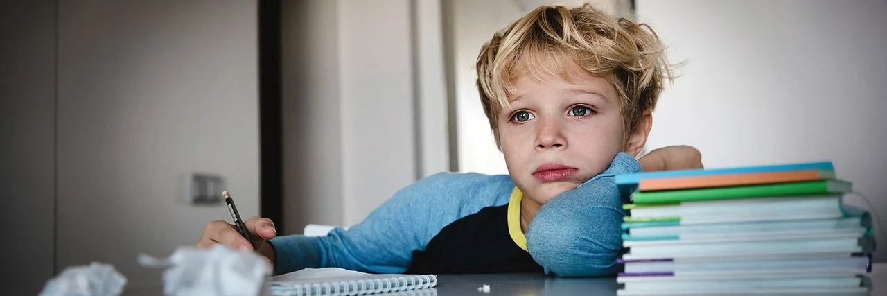 Ein Schuljunge sitzt an einem Tisch. Auf dem Tisch liegen ein Schreibblock, ein Stapel Lernmaterialien sowie zerknüllte Papierkugeln. In seiner rechten Hand hält er einen Stift. Er blickt nicht auf den Block, sondern nach vorne.