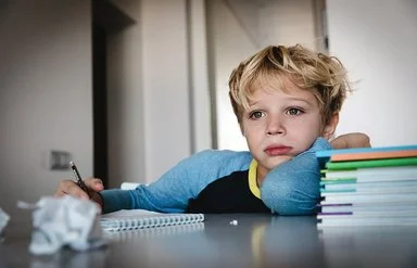 Ein Schuljunge sitzt an einem Tisch. Auf dem Tisch liegen ein Schreibblock, ein Stapel Lernmaterialien sowie zerknüllte Papierkugeln. In seiner rechten Hand hält er einen Stift. Er blickt nicht auf den Block, sondern nach vorne.