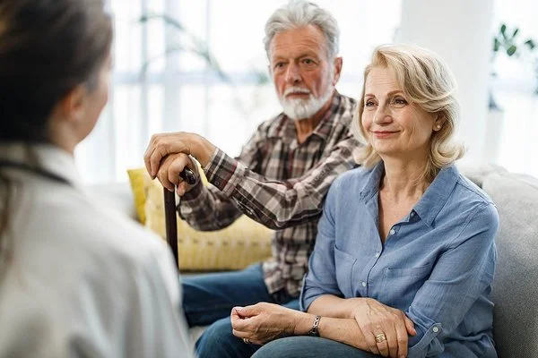 Ein älteres Paar sitzt in einem Wohnzimmer auf einer gemütlichen Couch. Die Frau wirkt entspannt, der Mann stützt seine Hände auf einem Gehstock vor ihm ab. Sie unterhalten sich mit einer Pflegerin, die ihnen gegenüber sitzt.