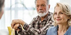 Ein älteres Paar sitzt in einem Wohnzimmer auf einer gemütlichen Couch. Die Frau wirkt entspannt, der Mann stützt seine Hände auf einem Gehstock vor ihm ab. Sie unterhalten sich mit einer Pflegerin, die ihnen gegenüber sitzt.