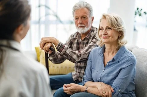 Ein älteres Paar sitzt in einem Wohnzimmer auf einer gemütlichen Couch. Die Frau wirkt entspannt, der Mann stützt seine Hände auf einem Gehstock vor ihm ab. Sie unterhalten sich mit einer Pflegerin, die ihnen gegenüber sitzt.