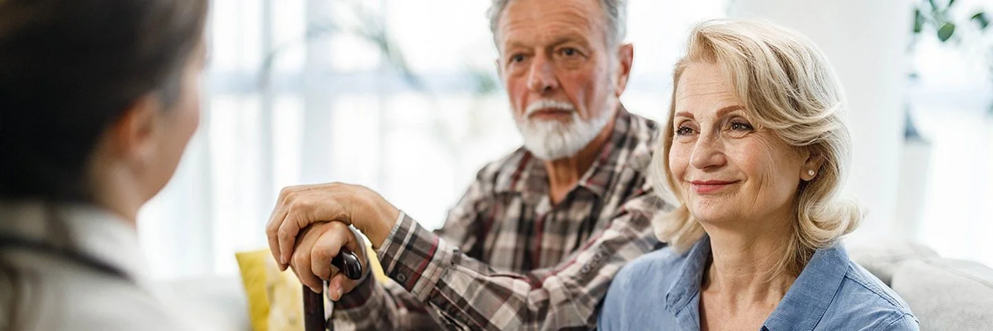Ein älteres Paar sitzt in einem Wohnzimmer auf einer gemütlichen Couch. Die Frau wirkt entspannt, der Mann stützt seine Hände auf einem Gehstock vor ihm ab. Sie unterhalten sich mit einer Pflegerin, die ihnen gegenüber sitzt.