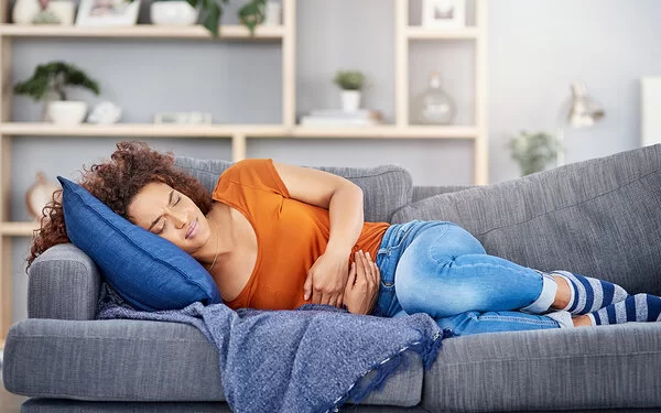 Eine junge Frau liegt mit schmerzverzerrtem Gesicht auf der Couch und hält sich den Bauch.