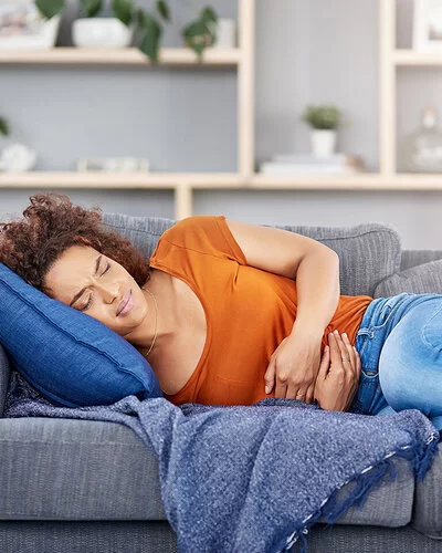 Eine junge Frau liegt mit schmerzverzerrtem Gesicht auf der Couch und hält sich den Bauch.