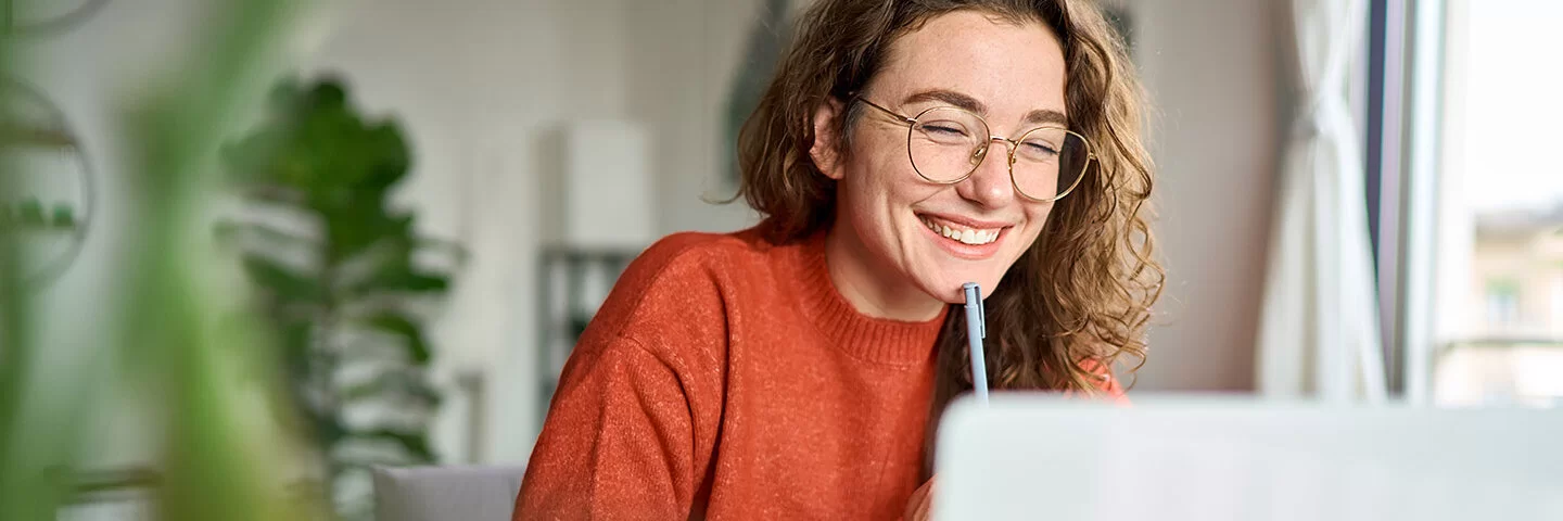 Eine junge Frau mit lockigen hellbraunen Haaren sitzt am Schreibtisch und schaut lächelnd auf ihren aufgeklappten Laptop. Sie trägt eine runde Brille mit dünnem Metallrahmen und einen dunkelorangen Pullover. Im Hintergrund ist ein Wohnzimmer mit heller Einrichtung und Grünpflanzen zu sehen.