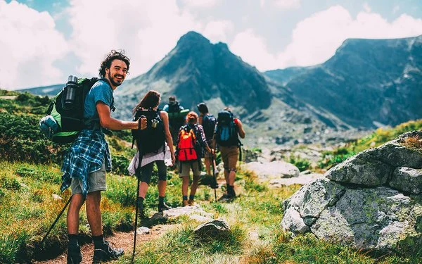 Eine Gruppe von sechs Menschen bewegt sich beim Trekking auf einem Pfad im Gebirge. Sie tragen alle vollgepackte große Rucksäcke und benutzen Trekkingstöcke. Die Gruppe ist von hinten zu sehen. Der Letzte in der Reihe, ein junger Mann, dreht sich um und lächelt in die Kamera.