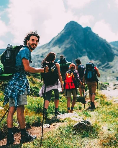 Eine Gruppe von sechs Menschen bewegt sich beim Trekking auf einem Pfad im Gebirge. Sie tragen alle vollgepackte große Rucksäcke und benutzen Trekkingstöcke. Die Gruppe ist von hinten zu sehen. Der Letzte in der Reihe, ein junger Mann, dreht sich um und lächelt in die Kamera.