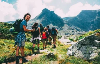 Eine Gruppe von sechs Menschen bewegt sich beim Trekking auf einem Pfad im Gebirge. Sie tragen alle vollgepackte große Rucksäcke und benutzen Trekkingstöcke. Die Gruppe ist von hinten zu sehen. Der Letzte in der Reihe, ein junger Mann, dreht sich um und lächelt in die Kamera.