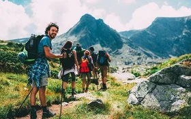 Eine Gruppe von sechs Menschen bewegt sich beim Trekking auf einem Pfad im Gebirge. Sie tragen alle vollgepackte große Rucksäcke und benutzen Trekkingstöcke. Die Gruppe ist von hinten zu sehen. Der Letzte in der Reihe, ein junger Mann, dreht sich um und lächelt in die Kamera.
