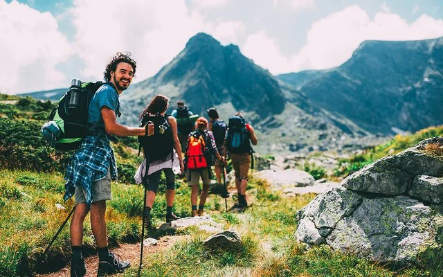 Eine Gruppe von sechs Menschen bewegt sich beim Trekking auf einem Pfad im Gebirge. Sie tragen alle vollgepackte große Rucksäcke und benutzen Trekkingstöcke. Die Gruppe ist von hinten zu sehen. Der Letzte in der Reihe, ein junger Mann, dreht sich um und lächelt in die Kamera.