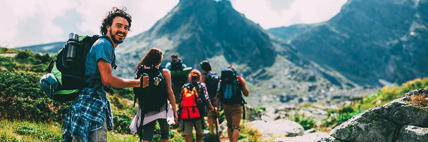Eine Gruppe von sechs Menschen bewegt sich beim Trekking auf einem Pfad im Gebirge. Sie tragen alle vollgepackte große Rucksäcke und benutzen Trekkingstöcke. Die Gruppe ist von hinten zu sehen. Der Letzte in der Reihe, ein junger Mann, dreht sich um und lächelt in die Kamera.