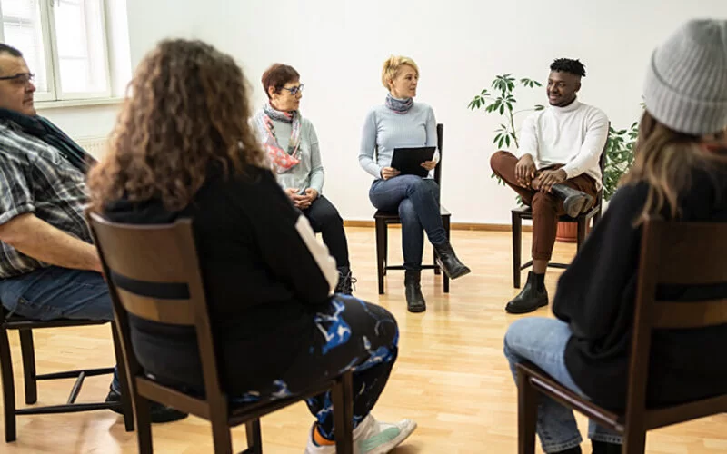 Eine Gruppe von sechs Menschen sitzt für eine Gruppen-Psychotherapie in einem hellen Raum im Stuhlkreis.