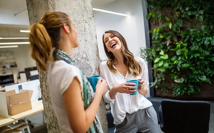 Zwei Frauen stehen in einem modernen Büro, eine lehnt an einer Säule, beide halten blaue Tassen und unterhalten sich.