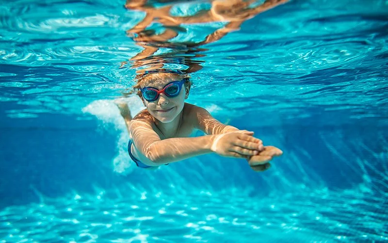 Junge im Grundschulalter taucht mit Schwimmbrille in einem Swimmingpool und lächelt.