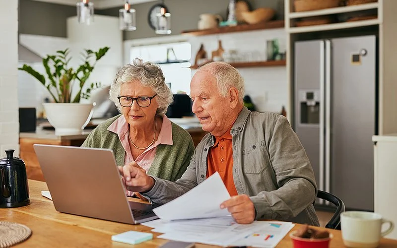 Ein älteres Ehepaar sitzt am Küchentisch vor einem Laptop. Neben dem Mann liegen Papiere auf dem Tisch. Ein Blatt hält er in der Hand.