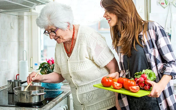 Eine alte Frau steht in der Küche und schwenkt einen Löffel im Kochtopf, während eine jüngere Frau daneben steht und zuguckt.