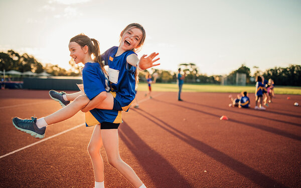 Zwei Mädchen gehen in Sportkleidung im Abendlicht über einen Sportplatz. Das eine trägt das andere Huckepack.