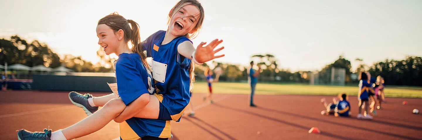 Zwei Mädchen gehen in Sportkleidung im Abendlicht über einen Sportplatz. Das eine trägt das andere Huckepack.