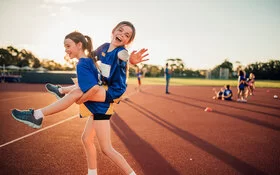 Zwei Mädchen gehen in Sportkleidung im Abendlicht über einen Sportplatz. Das eine trägt das andere Huckepack.