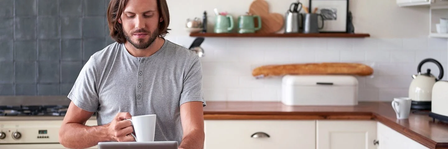 Ein Mann im grauen T-Shirt steht mit einer Tasse in der Hand vor dem Küchentresen und schaut auf sein Tablet.
