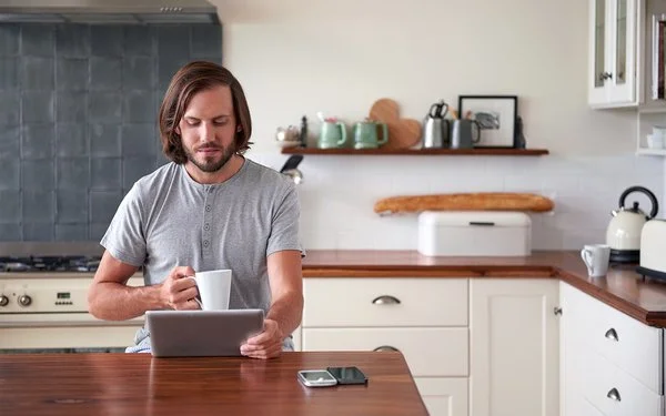 Ein Mann im grauen T-Shirt steht mit einer Tasse in der Hand vor dem Küchentresen und schaut auf sein Tablet.