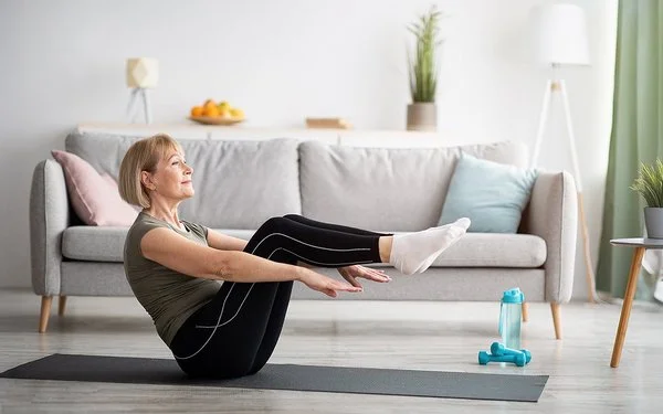 Eine ältere Frau in Sportkleidung macht auf einer Yoga-Matte in einem Wohnzimmer die halbe Boot-Übung.