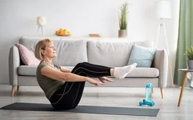 Eine ältere Frau in Sportkleidung macht auf einer Yoga-Matte in einem Wohnzimmer die halbe Boot-Übung.