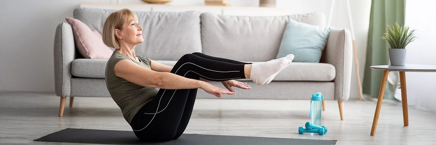 Eine ältere Frau in Sportkleidung macht auf einer Yoga-Matte in einem Wohnzimmer die halbe Boot-Übung.