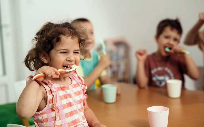 Drei Kinder sitzen an einem Tisch und putzen sich gemeinsam mit bunten Zahnbürsten die Zähne. Das Mädchen im Vordergrund lacht fröhlich.