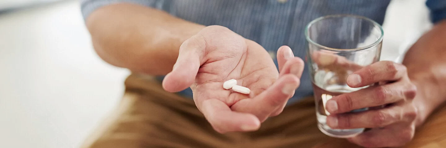 Ein Mann hält zwei Tabletten in der Hand, die er mit einem Glas Wasser zu sich nehmen möchte.