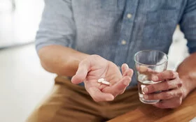 Ein Mann hält zwei Tabletten in der Hand, die er mit einem Glas Wasser zu sich nehmen möchte.