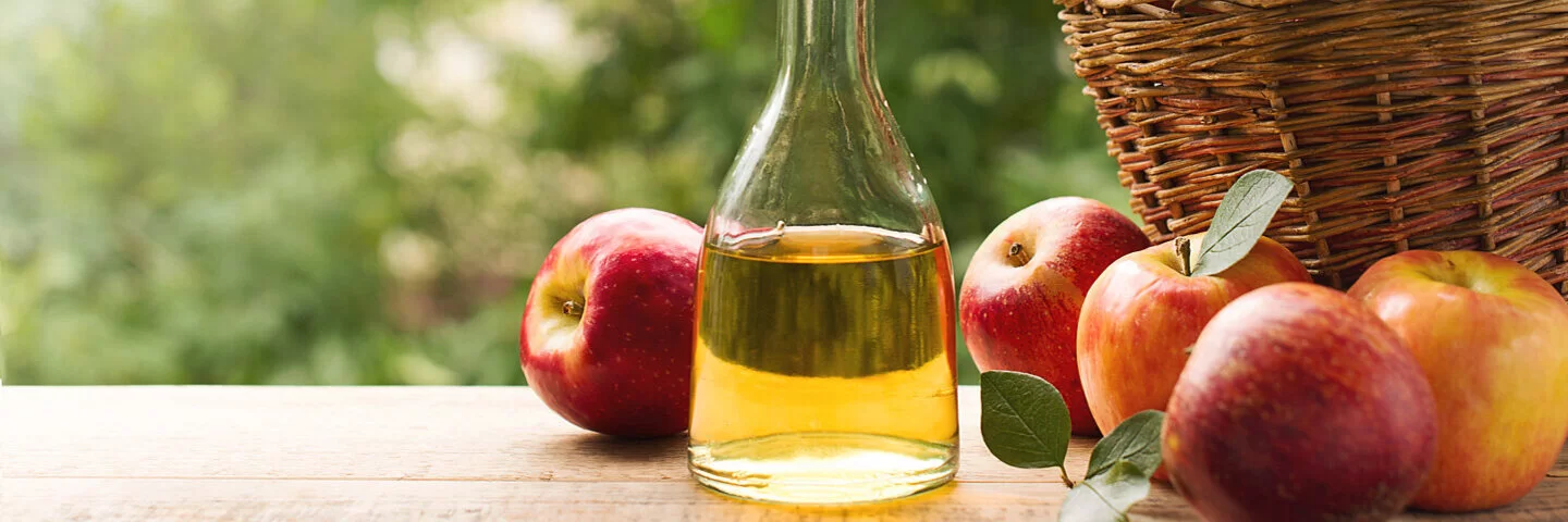 Eine Flasche mit Apfelessig neben einem Korb mit Äpfeln.